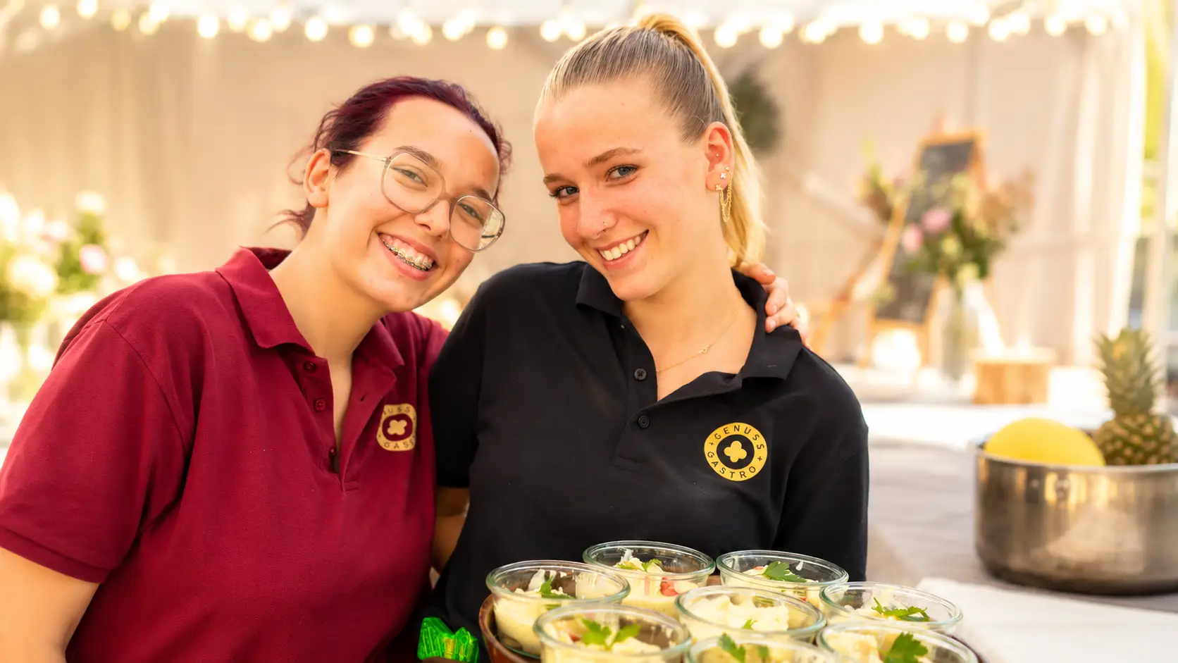 Zwei lachende Mitarbeiterinnen im weinroten und schwarzen Polo tragen gemeinsam ein Tablett mit Salatgläschen