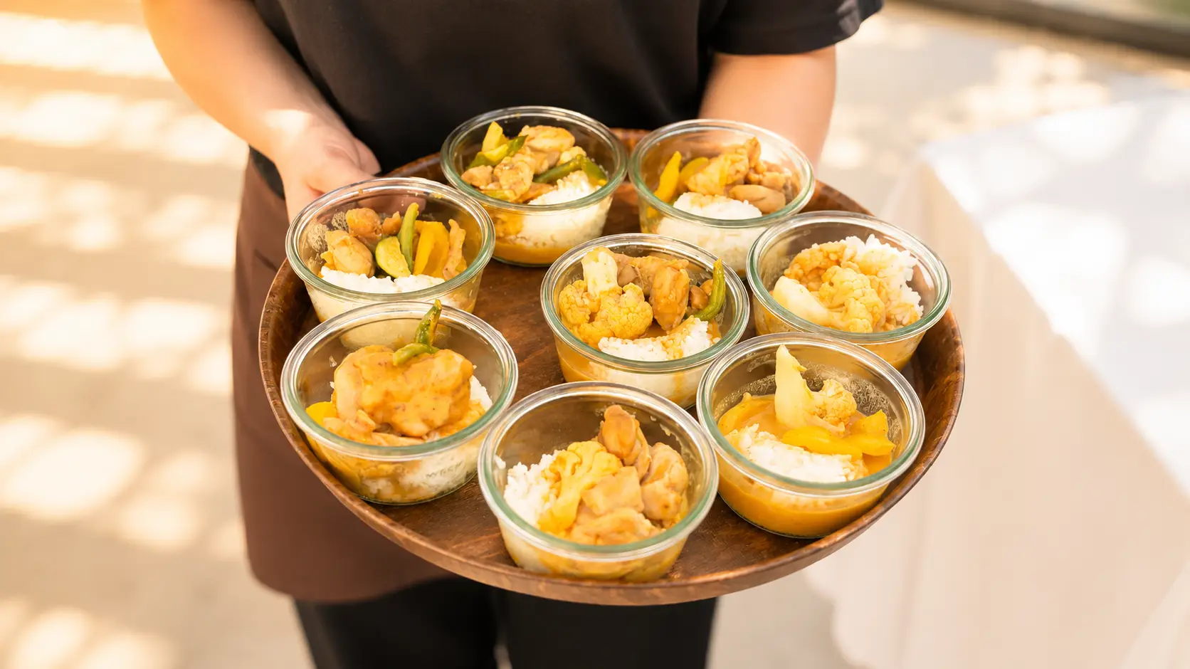 Rundes Holztablett mit Gläschen voller Reis, Curry und Ananas, von einer Servicekraft getragen