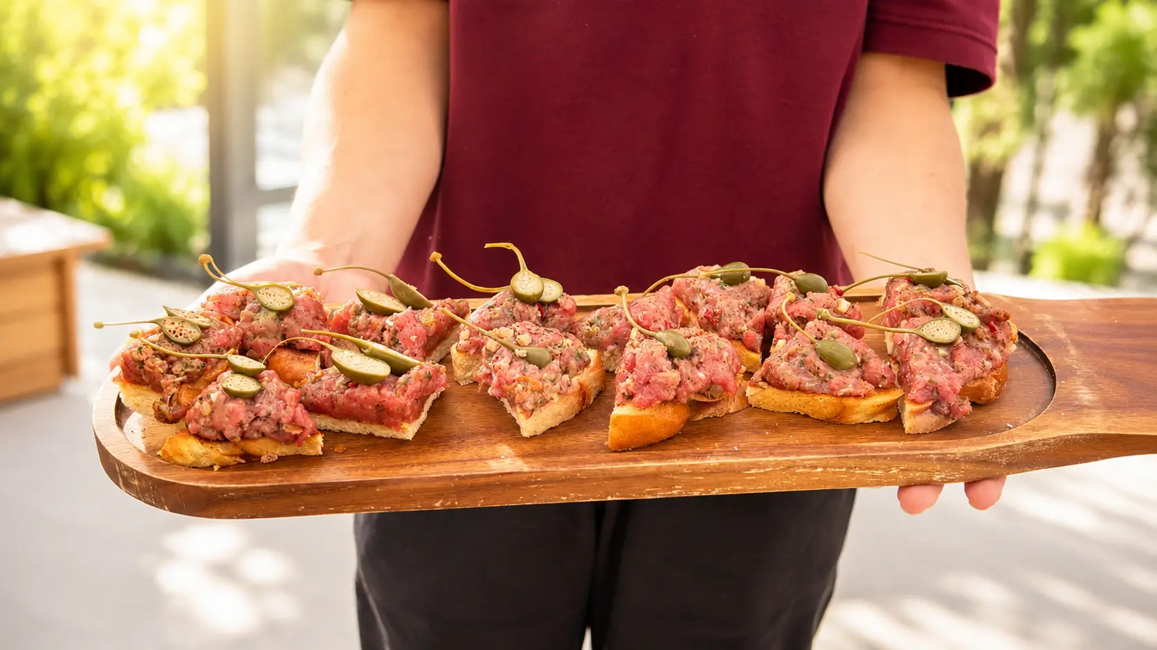 Langes Holzbrett mit Crostini, belegt mit Tatar und Gurken, von einer Servicekraft im Freien gehalten