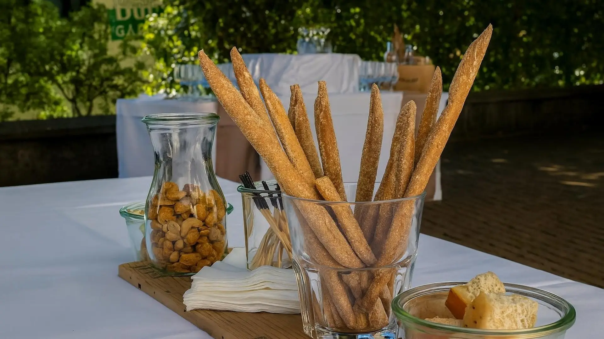 Holzbrett auf einem weiss gedeckten Tisch im Freien mit Grissini im Glas, Nüssen und Käsewürfeln