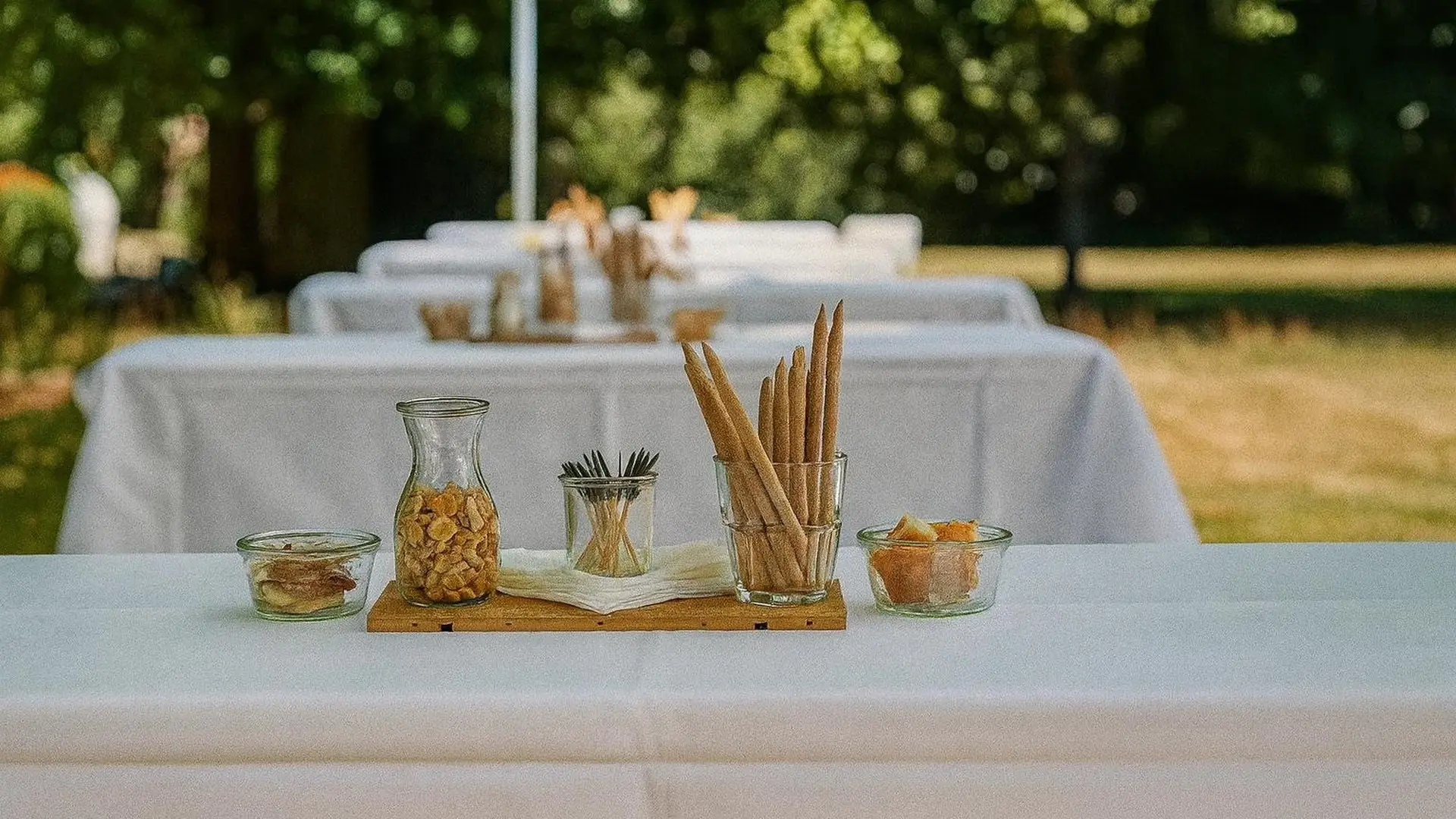 Reihe weiss gedeckter Apéro-Tische im Park, darauf Holzbretter mit Grissini, Nüssen und Knabbereien im Glas