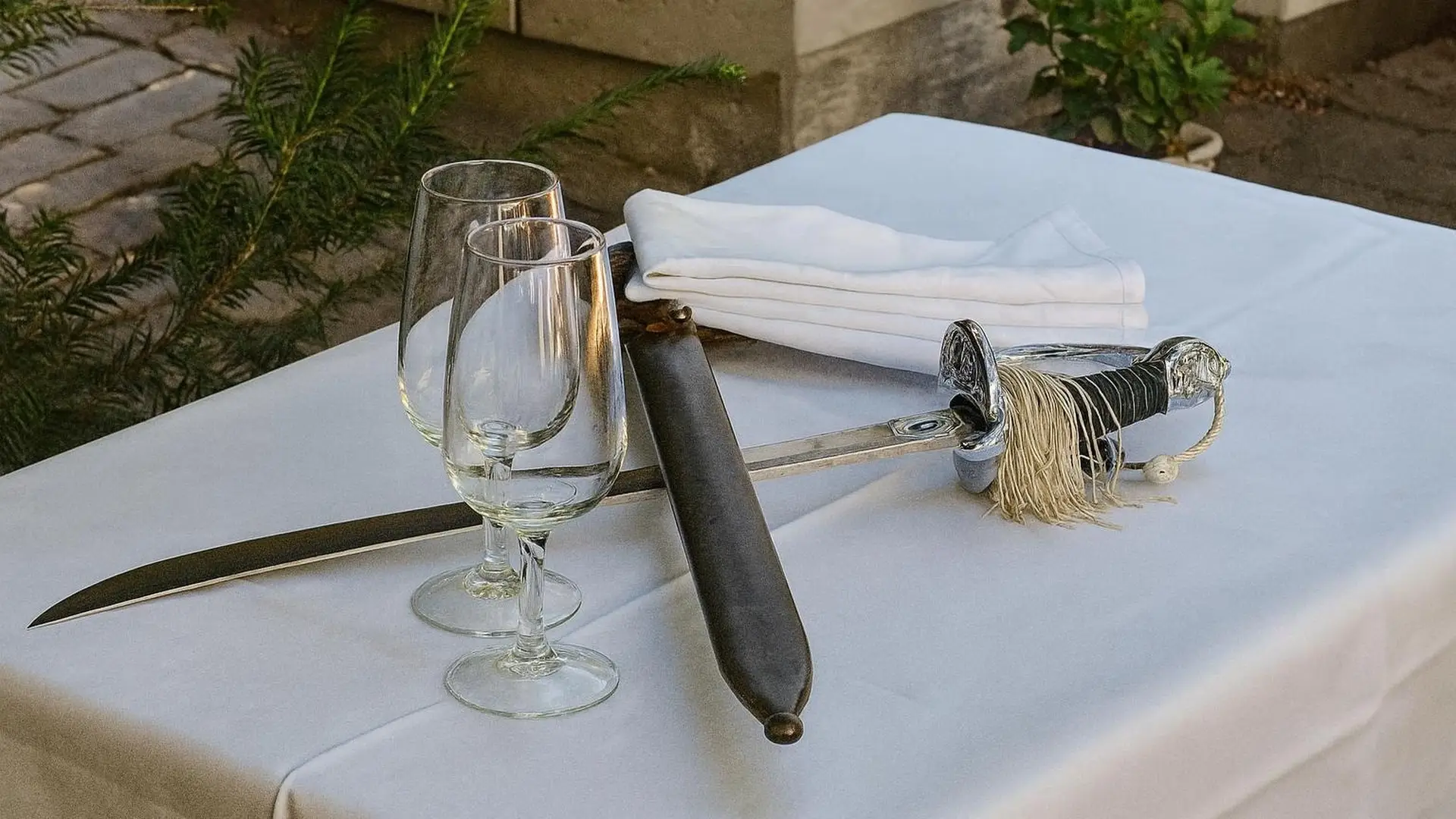 Weiss gedeckter Tisch im Freien mit zwei Sektgläsern, einer weissen Serviette und einem Säbel zum Köpfen der Flasche