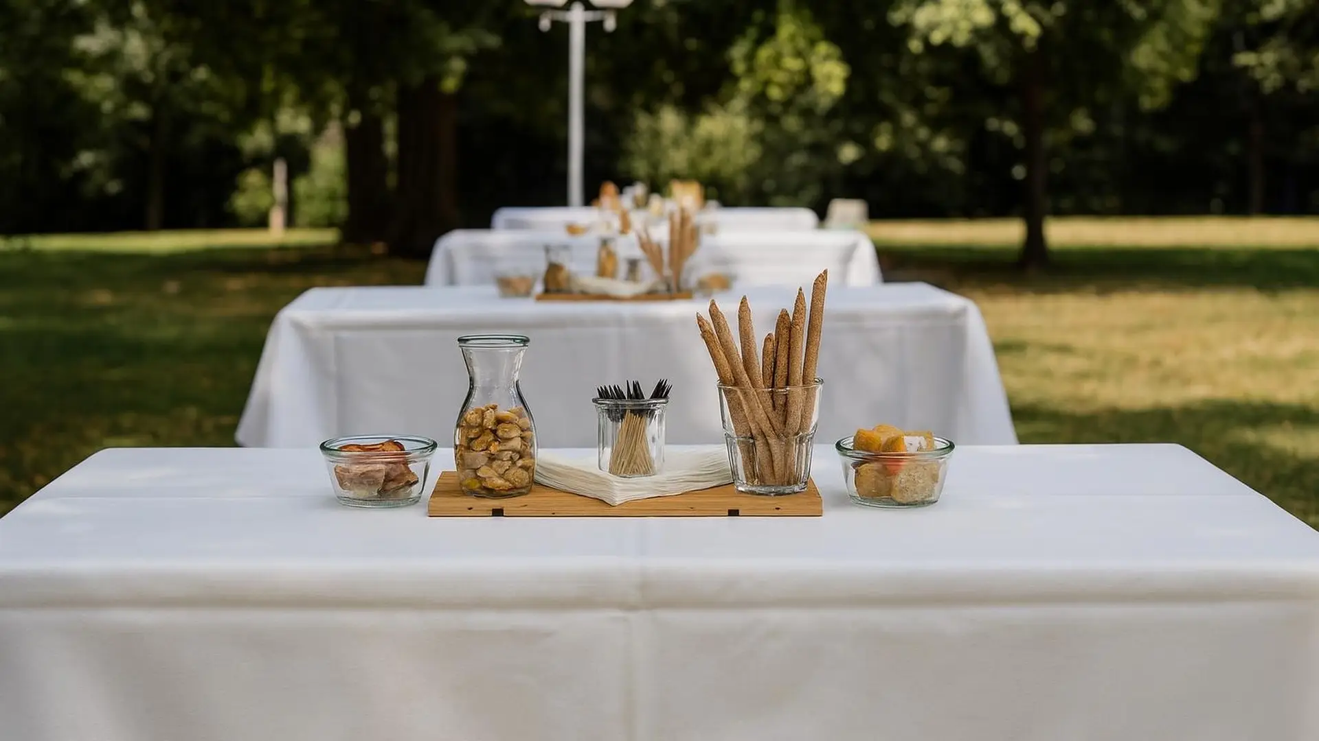 Weiss gedeckte Tische auf einer Wiese im Park, darauf Holzbretter mit Grissini, Nüssen und Knabbereien im Glas
