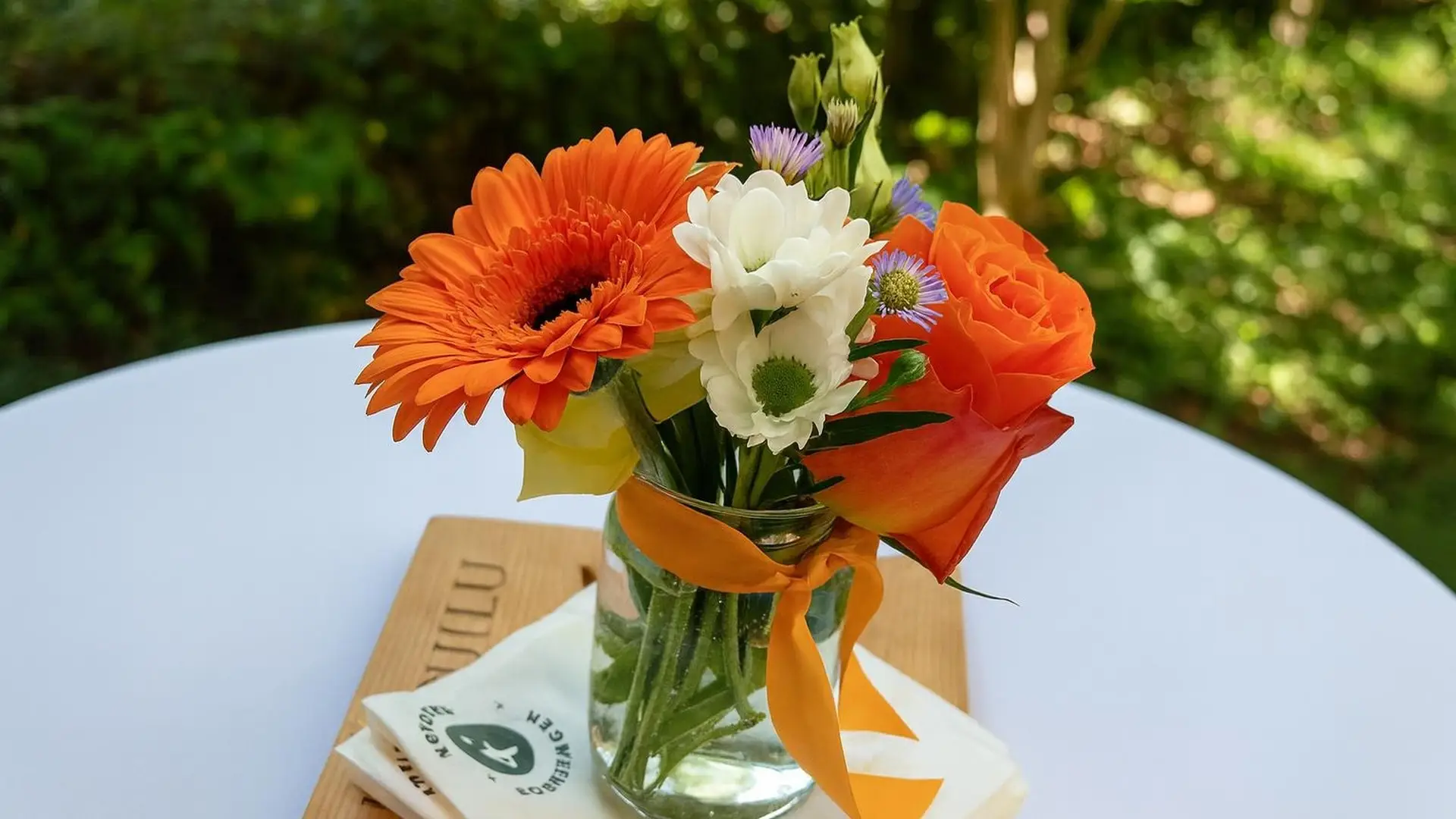 Glasvase mit orangen Gerbera, weissen Blüten und einer Rose auf einem weiss gedeckten Tisch im Grünen, daneben eine Karte