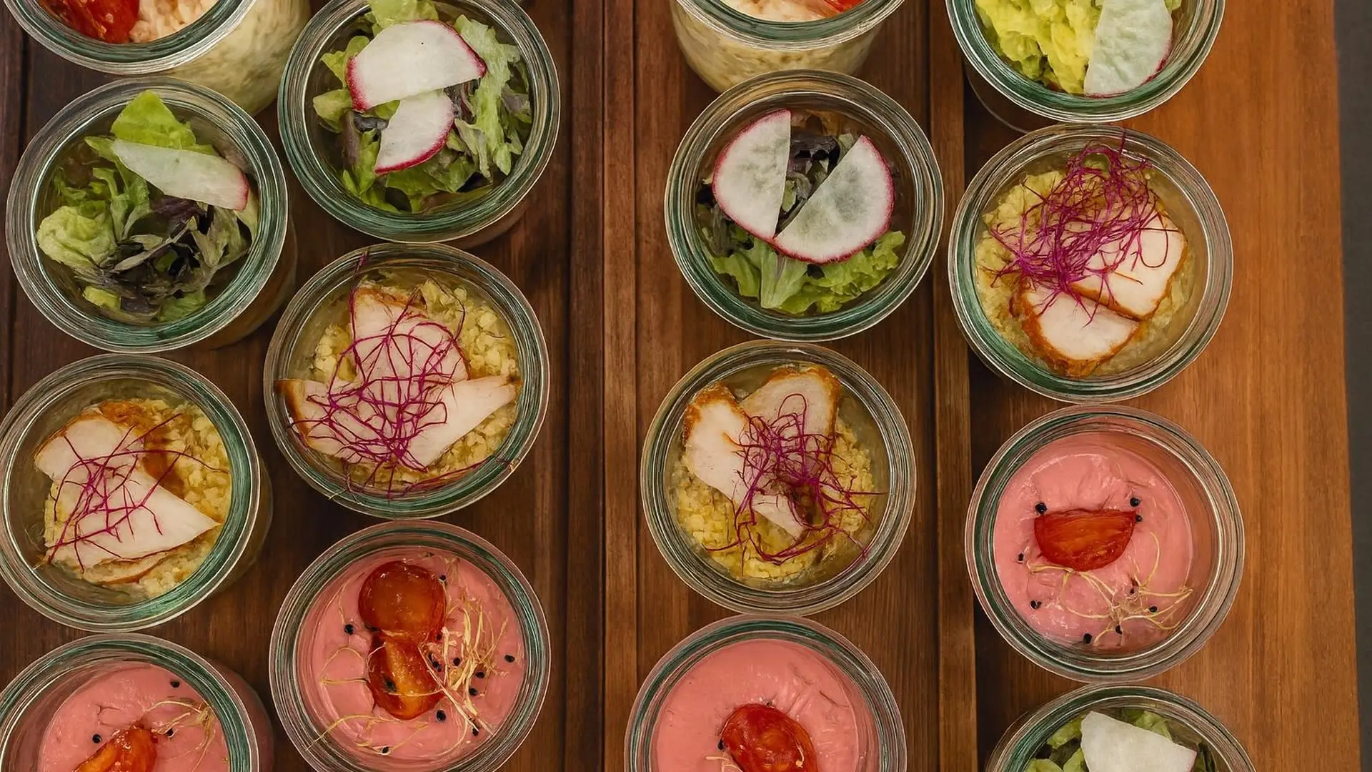 Blick von oben auf Holzbretter mit Gläschen voller Blattsalat, Couscous, rosa Creme und Radieschen