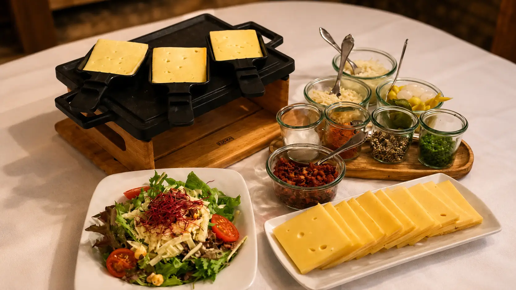 Raclette-Ofen mit Käsescheiben auf weiss gedecktem Tisch, dazu ein Salatteller, eine Käseplatte und Beilagen in kleinen Gläsern