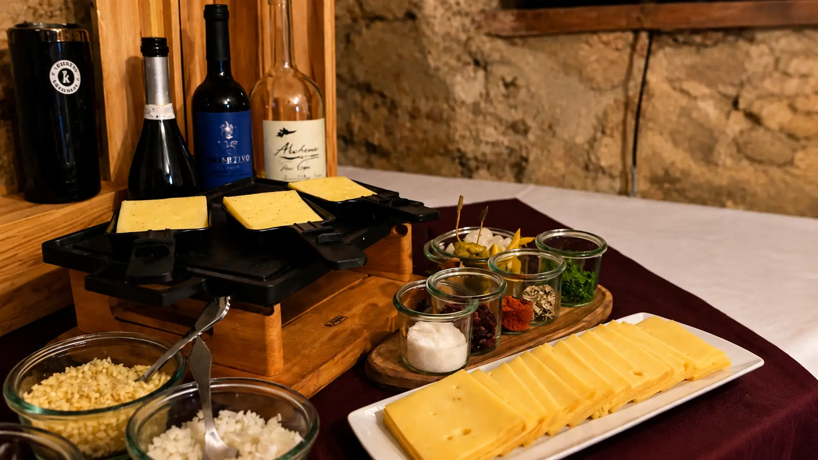 Raclette-Ofen mit Käsescheiben vor einer Bruchsteinwand, dazu eine Käseplatte, Beilagen in Gläsern und Weinflaschen