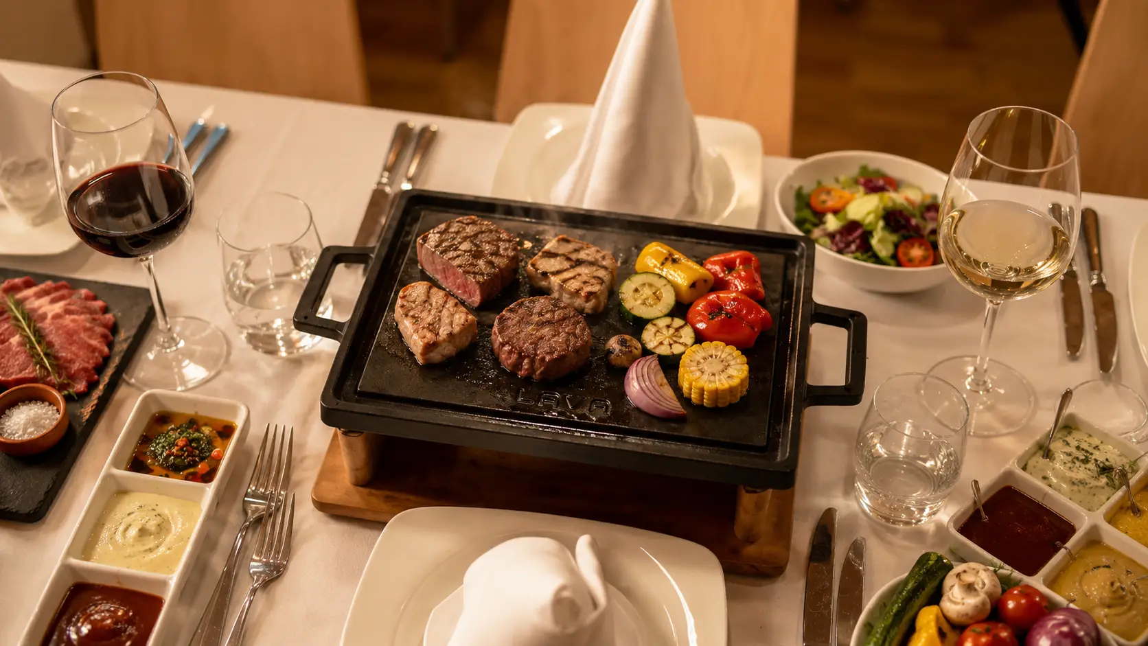 Tischgrill mit Steaks, Maisstücken und Gemüse auf einem gedeckten Tisch, dazu Saucen in kleinen Schälchen, Salat, Rot- und Weisswein