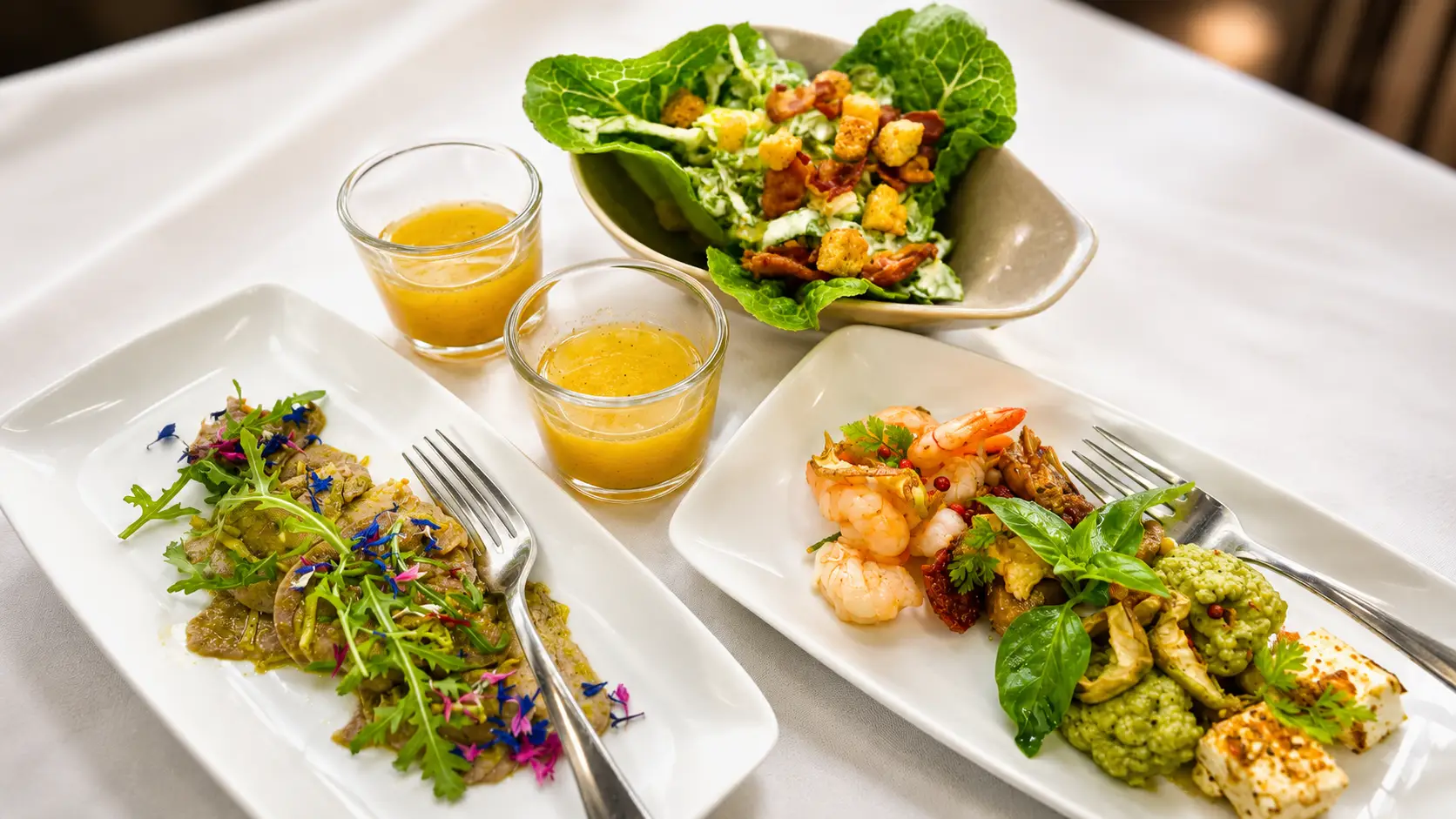 Weisse Teller mit Salat im Blattkörbchen, zwei Suppengläschen, Crevetten und Salat mit essbaren Blüten