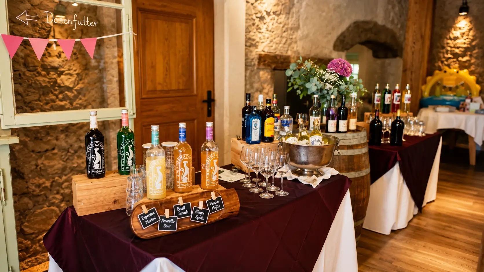 Aufgebaute Bar mit Spirituosen- und Weinflaschen, Schiefertäfelchen, Weingläsern und Sektkübel vor Weinfässern und Bruchsteinwand