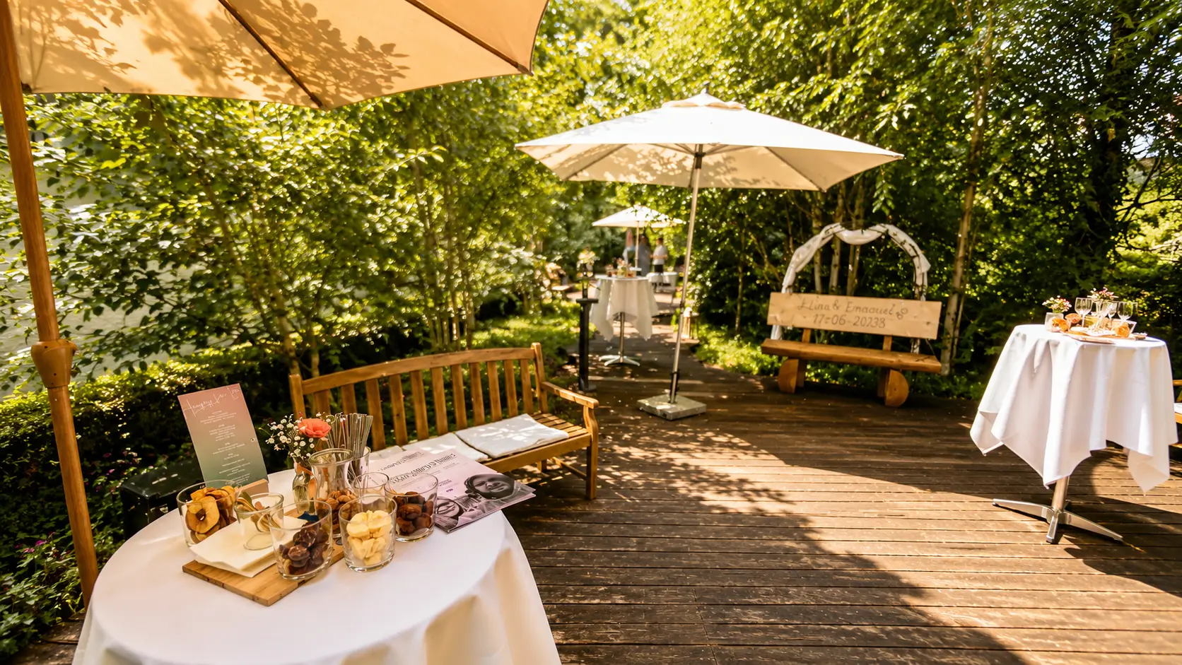 Stehtisch mit weisser Husse auf einer Holzterrasse im Grünen, darauf Knabbereien im Glas und eine Menükarte, darüber ein Sonnenschirm
