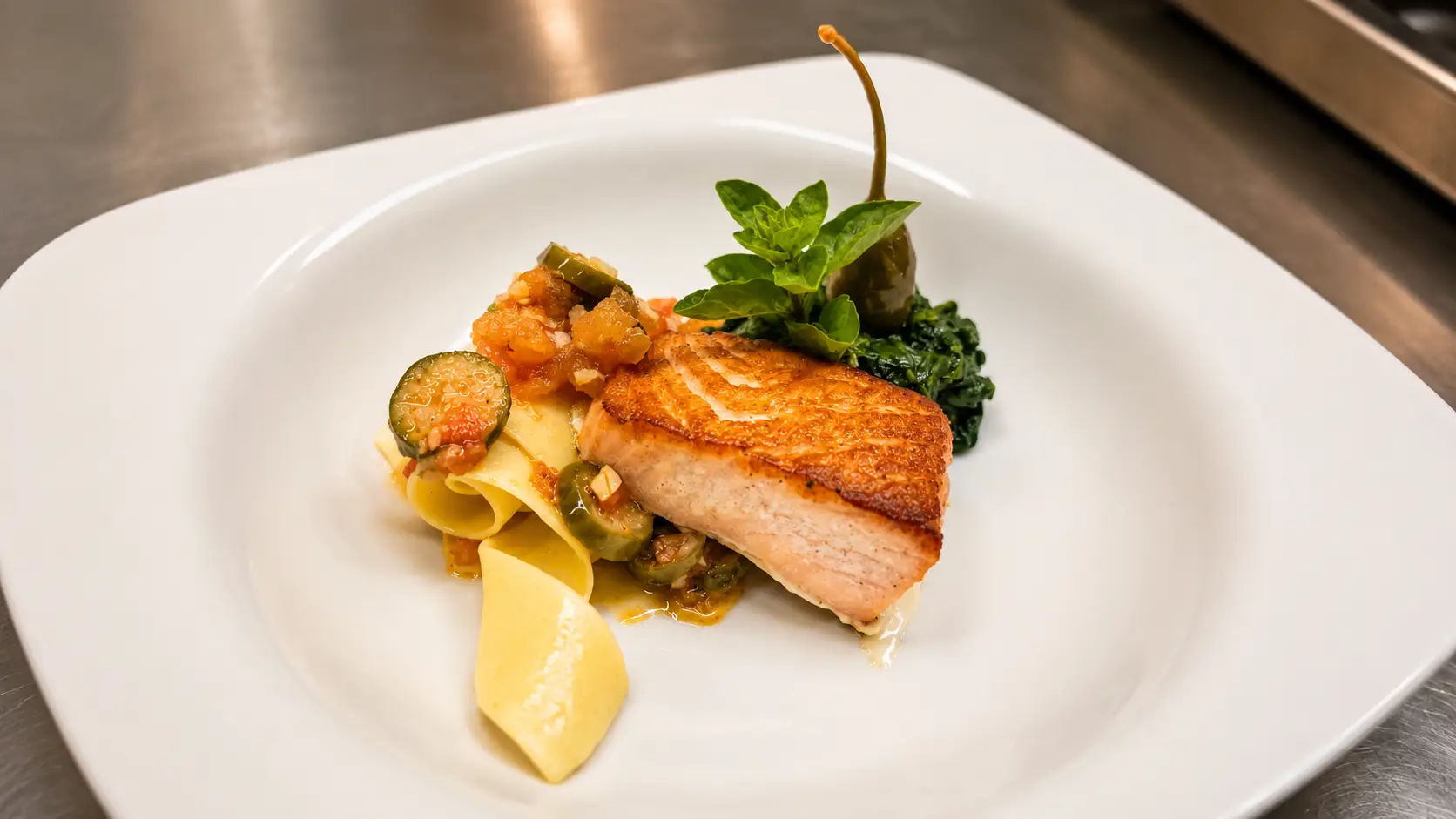Gebratenes Lachsfilet mit knuspriger Haut auf weissem Teller, dazu Zucchetti, Tomatengemüse, Kartoffeln und ein Kräuterzweig