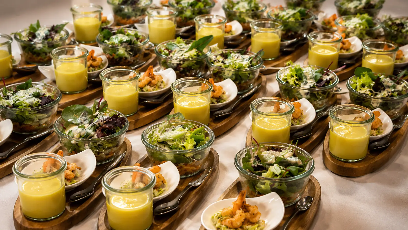 Holzbretter mit Reihen von Salatgläschen, gelben Suppengläschen und kleinen Tellern mit Crevetten