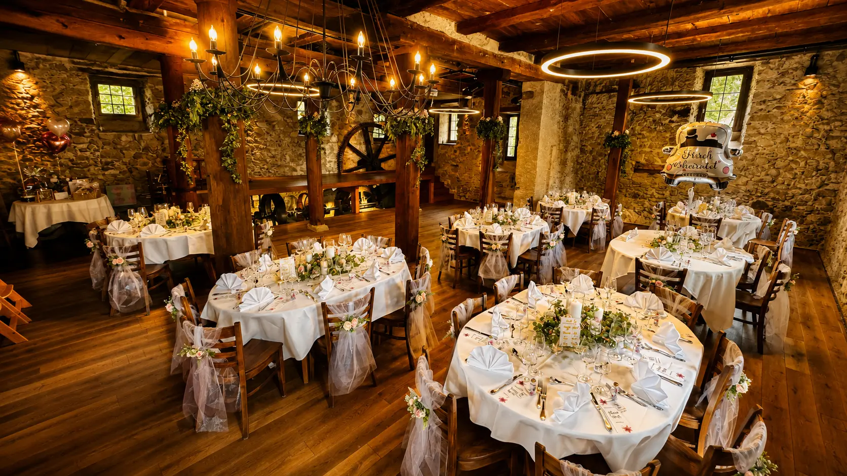Saal mit Bruchsteinwänden, Holzbalken und altem Mühlrad, darin runde eingedeckte Tische mit Stuhlhussen und Blumenschmuck