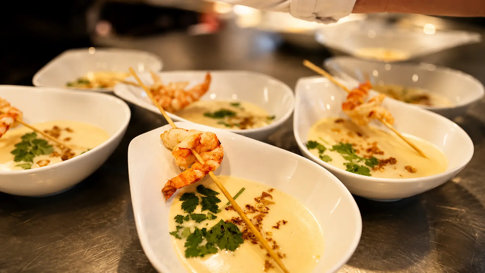 Flache Schalen mit heller Rahmsuppe, Kräuteröl und einem Spiess mit Crevetten, in der Küche zum Servieren bereitgestellt
