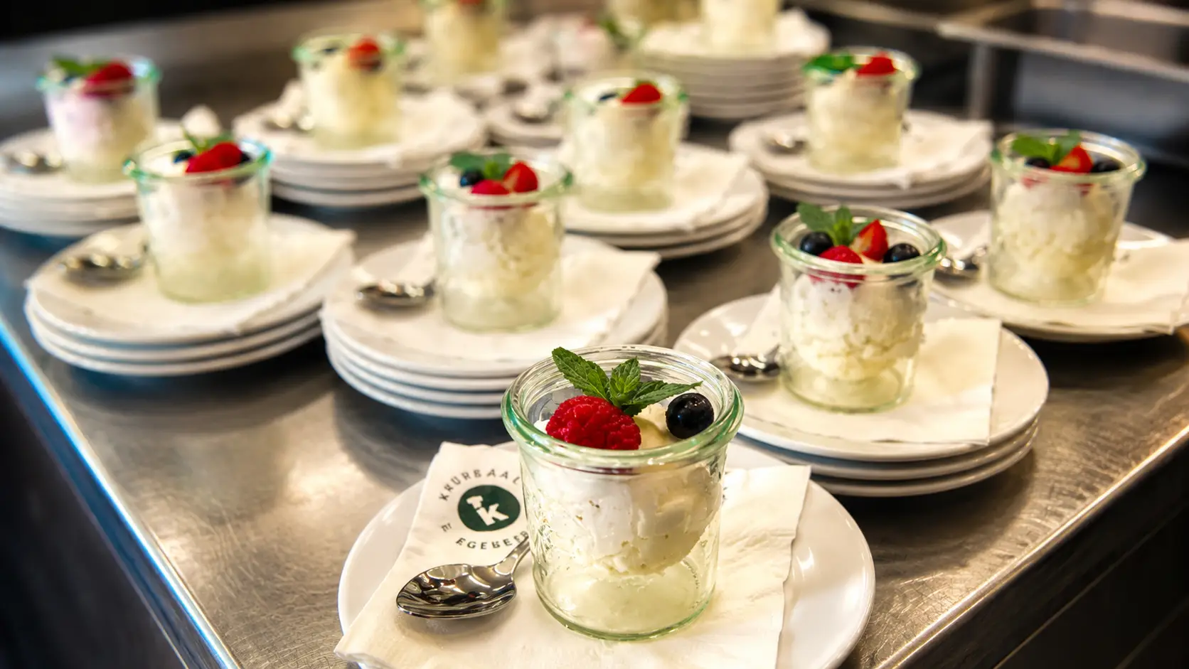 Dessertgläschen mit heller Creme, Himbeere, Brombeere und Minze, auf Tellerstapeln in der Küche zum Servieren bereitgestellt