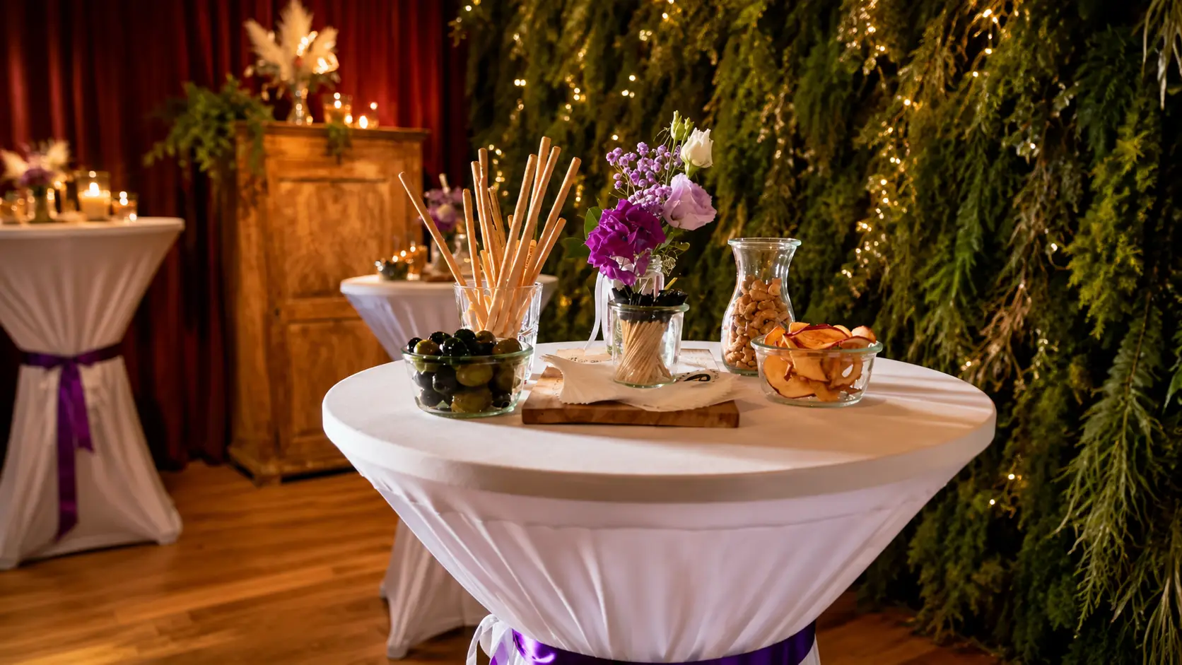 Stehtisch mit weisser Husse und lila Band vor einer Tannenwand mit Lichterketten, darauf Oliven, Grissini, Chips und Blumen