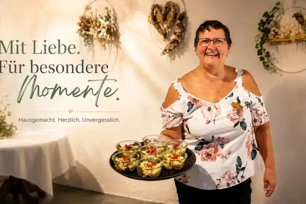 Monique Stampfler in geblümter Bluse trägt lachend ein Tablett mit Apéro-Gläschen vor einer Wand mit Trockenblumenkränzen; Bildtext «Mit Liebe. Für besondere Momente.»