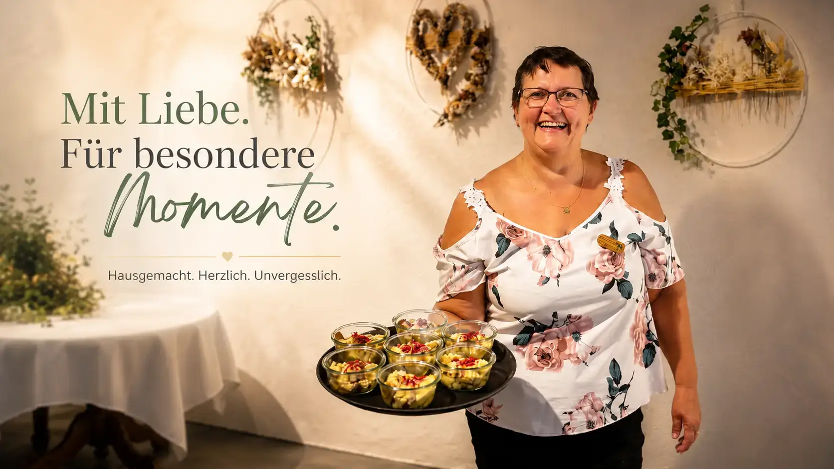 Monique Stampfler in geblümter Bluse trägt lachend ein Tablett mit Apéro-Gläschen vor einer Wand mit Trockenblumenkränzen